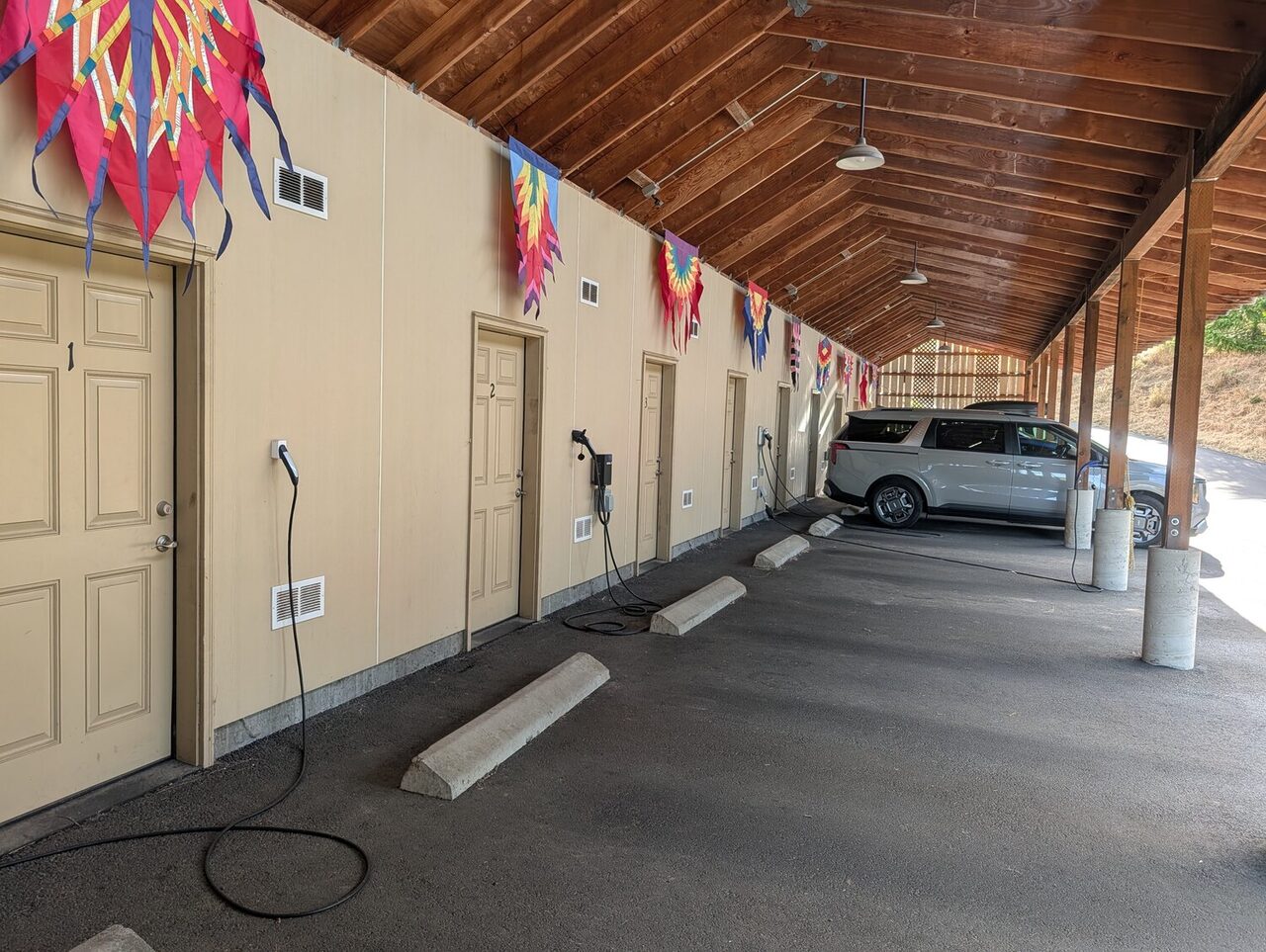 A row of numbered storage-unit doors under a timber roof, an EV charger beside each one and wheel stops marking the assigned parking spaces.