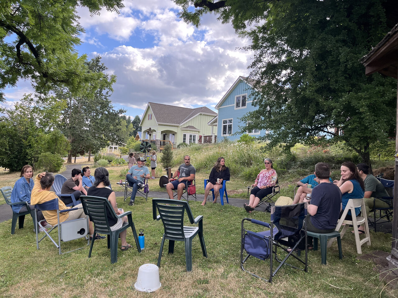 Neighbors sitting in a wide circle of lawn chairs on the grass, houses and trees behind them.