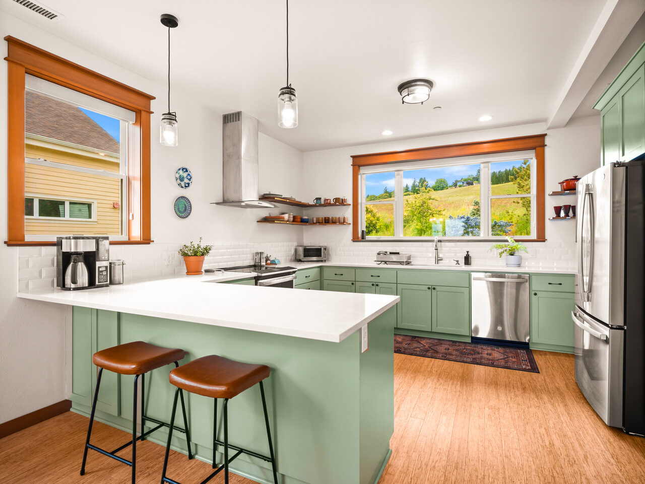 The kitchen: green cabinets, a white quartz island with two stools, an induction range under a hood, and a window over the sink looking out to fields and trees.