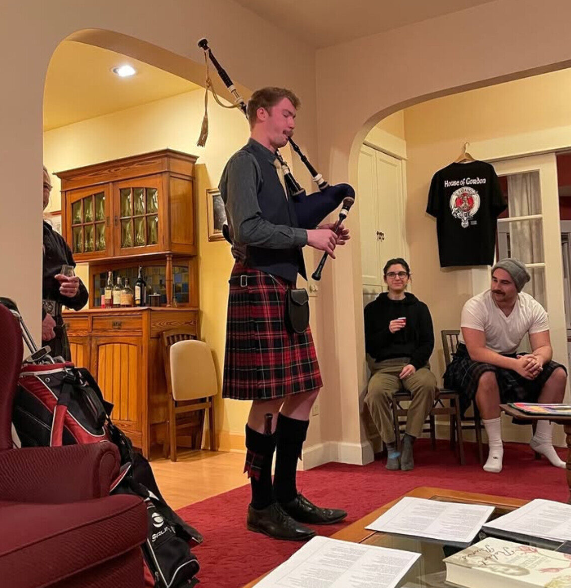 A piper in full kilt playing the bagpipes indoors at the Common House.